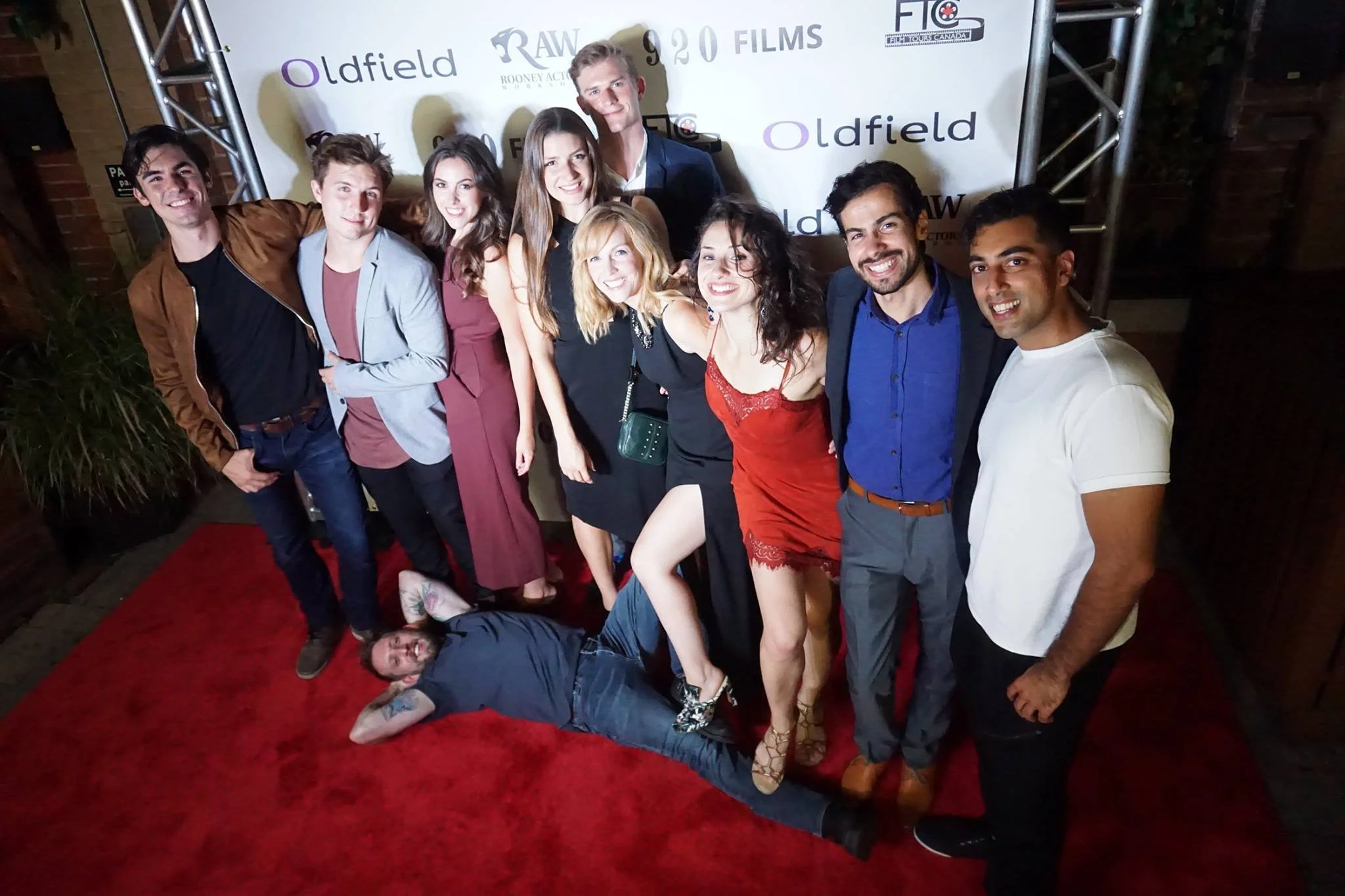 A group of eleven young people posing together on a red carpet at an event, with a backdrop featuring logos and sponsor names. They are smiling and dressed in semi-formal to stylish casual attire. One person is lying on the ground in front of the group.