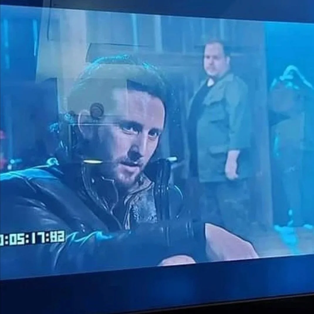 A scene from a movie or TV show featuring a man with dark hair and beard wearing a jacket, sitting at a table with a serious expression, with another man standing in the background in a room with wooden shelves and clothing.