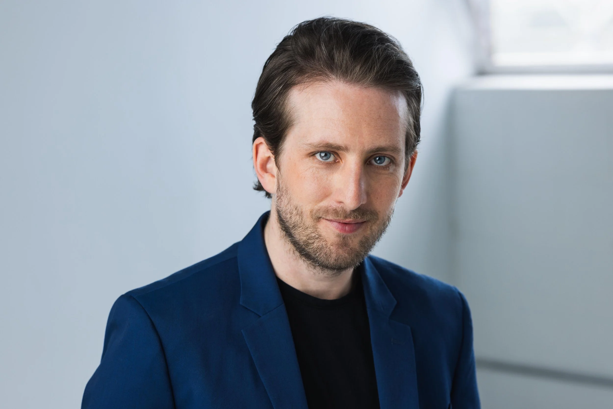 Portrait of a man, Michael Joseph Delaney, with brown hair and blue eyes wearing a dark blue blazer, smiling slightly, with a light background.