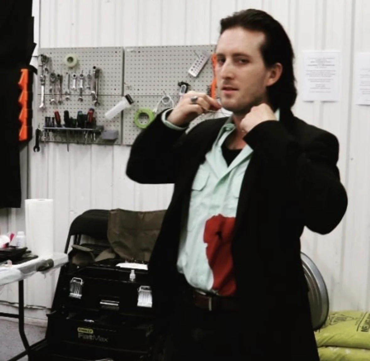 A man with dark hair and a black blazer standing in a workshop or garage, adjusting his collar, with tools hanging on a pegboard behind him.