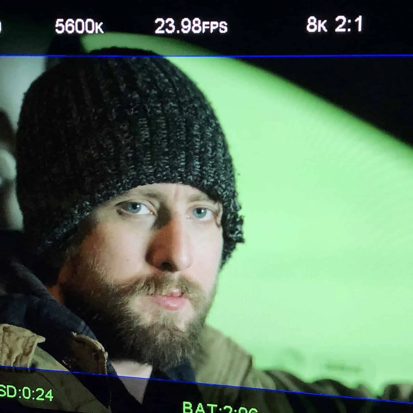 Close-up shot of a man with a beard and blue eyes, wearing a dark knitted beanie, with a green background.