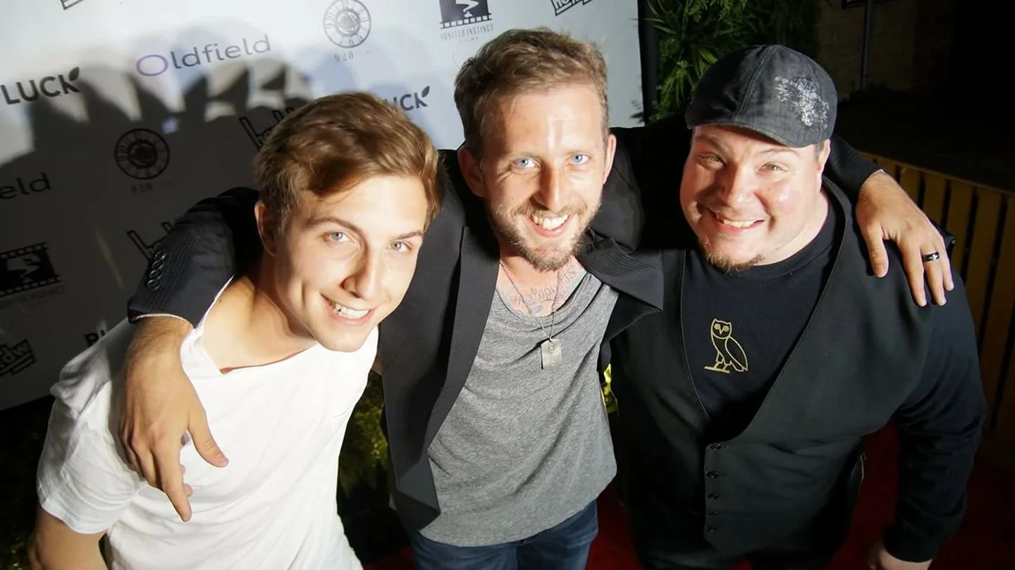 Three men are smiling and posing closely together. The man on the left has light brown hair and is wearing a white T-shirt. The man in the middle has light brown hair, a beard, and is wearing a gray shirt with a black jacket. The man on the right is wearing a black baseball cap and a black jacket with a yellow owl logo, and has his arm around the middle man's shoulder. They are standing in front of a backdrop with various logos.