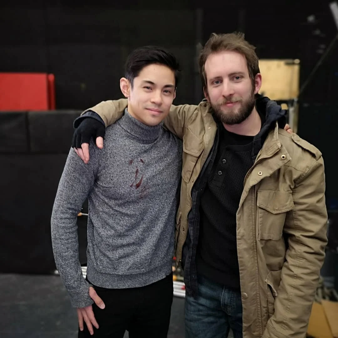Two young men standing close together, with one putting his arm around the other's shoulder, smiling at the camera in an indoor setting.