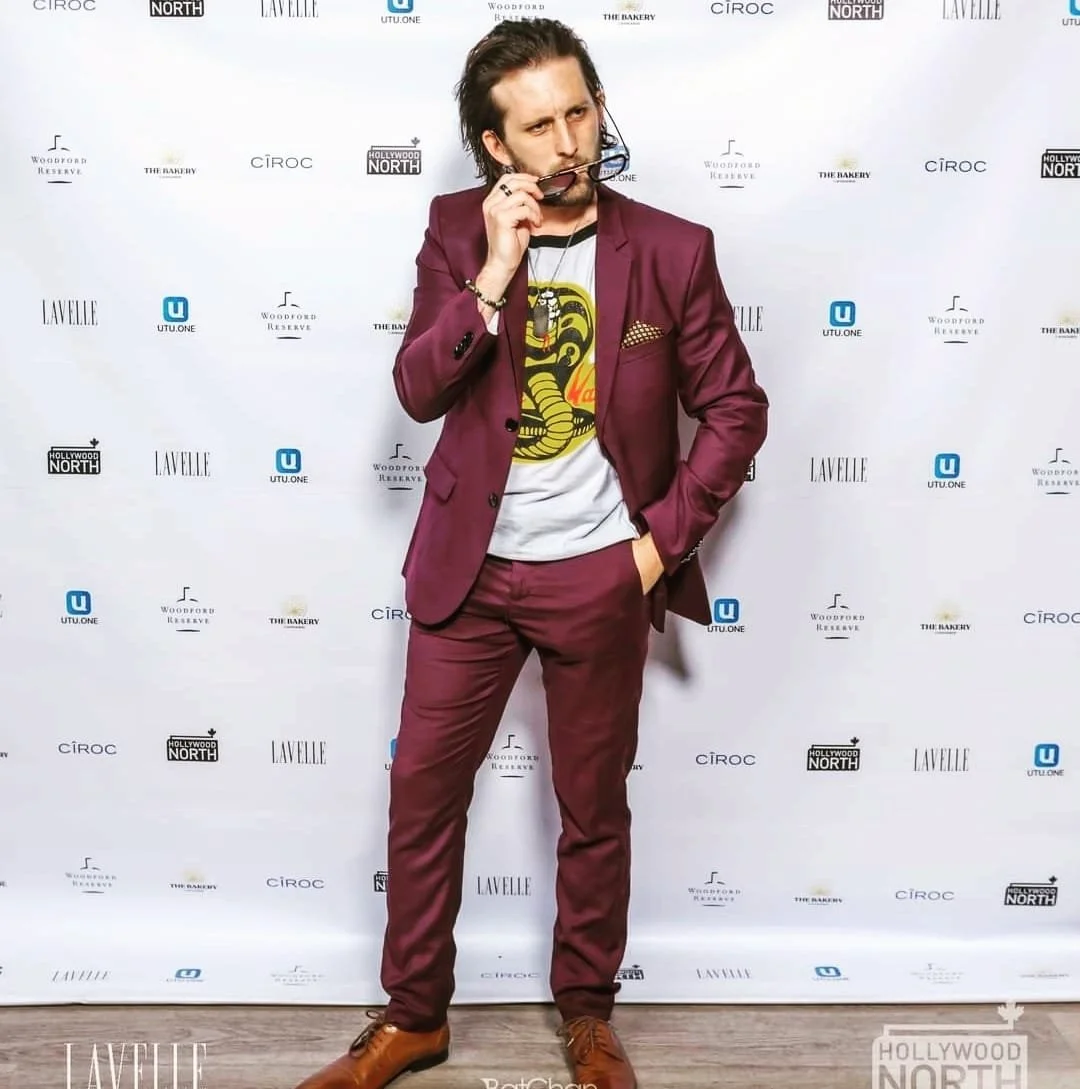 A man in a burgundy suit and brown shoes is standing in front of a step-and-repeat backdrop holding sunglasses and looking at the camera, with long dark hair.