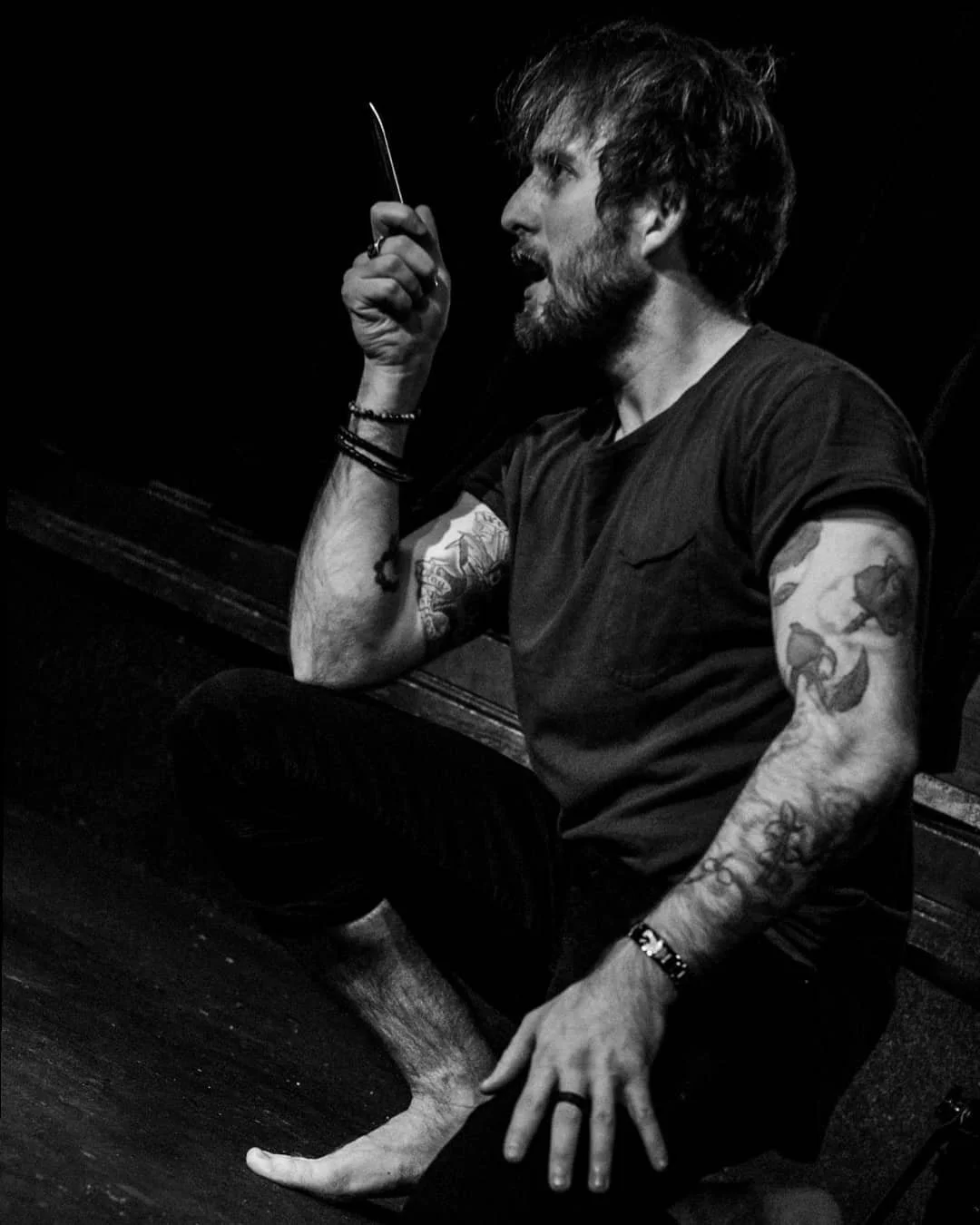 Black and white photo of a man sitting on a bench with one foot on the ground and the other foot on the bench. He has tattoos on both arms, a beard, and messy hair. He is holding a cigarette in one hand and appears to be smoking or in a moment of reflection.