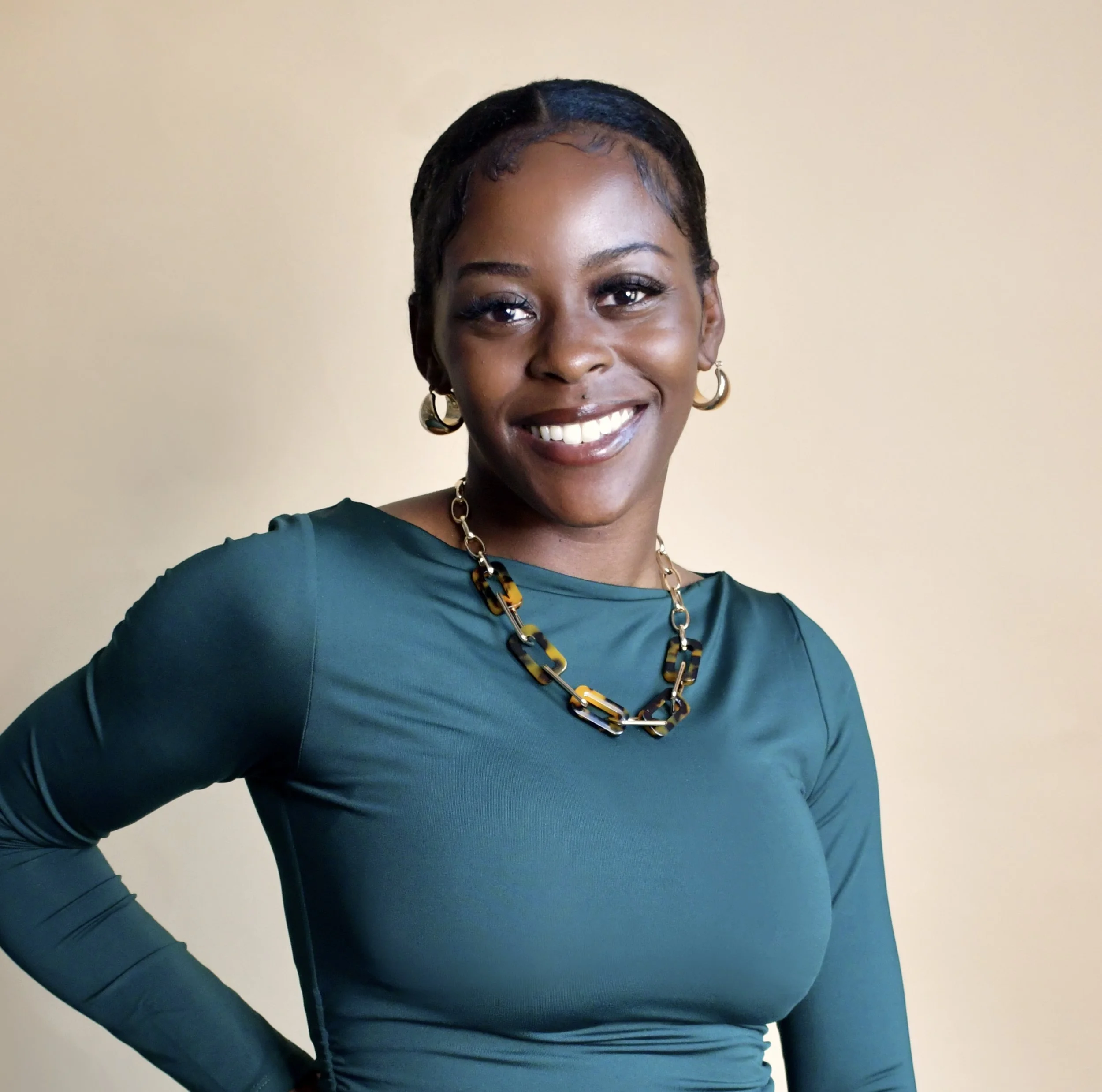 A smiling African American woman wearing a teal long-sleeve top, gold hoop earrings, a gold chain necklace with black and brown links, with her hand on her hip against a plain light-colored wall.