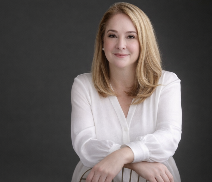 A woman with blonde hair in a white blouse, sitting with her arms crossed, smiling softly against a gray background.