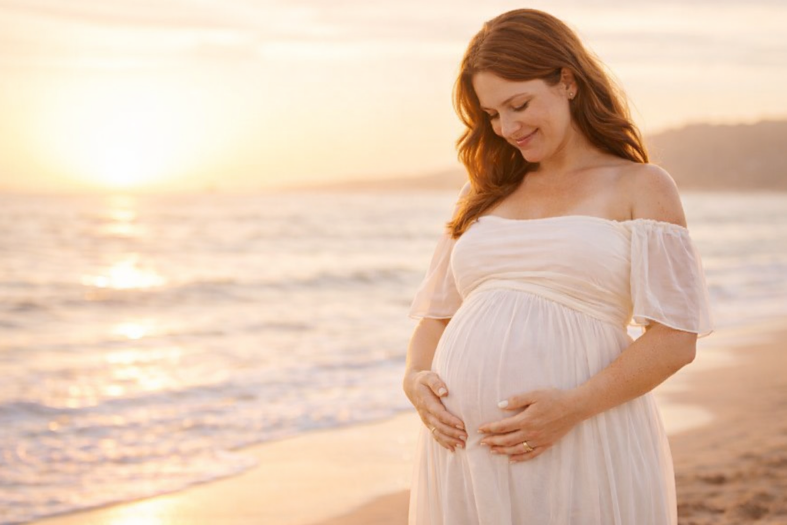 A pregnant woman with long red hair stands on a beach during sunset, gently holding her baby bump with both hands and smiling softly.
