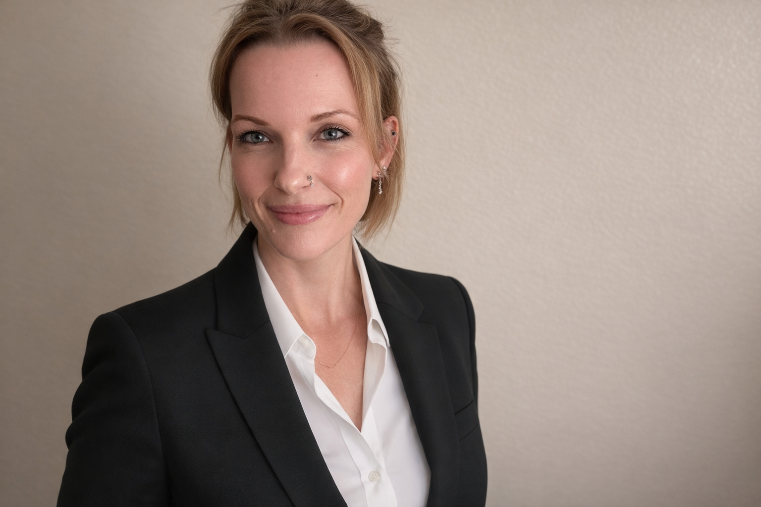 Portrait of a woman with short blonde hair, wearing a black blazer and white shirt, smiling and standing against a plain beige wall.