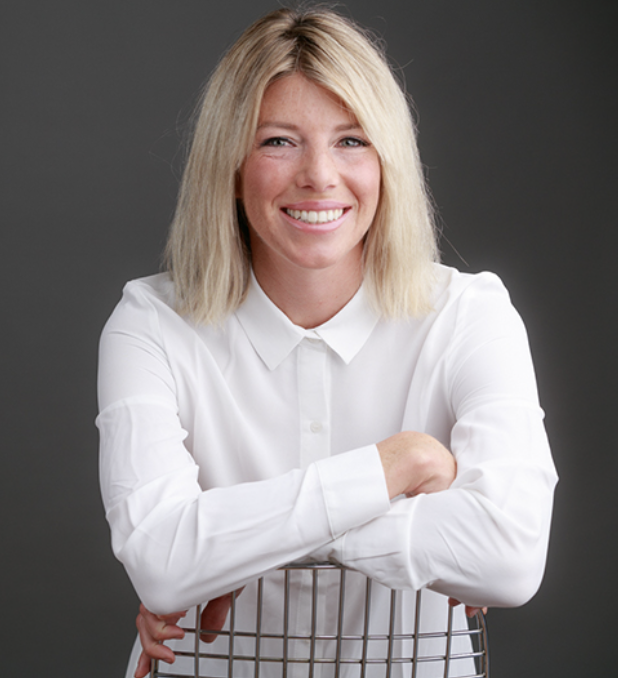 A woman with shoulder-length blonde hair wearing a white button-up shirt, smiling and leaning on a shopping cart against a gray background.
