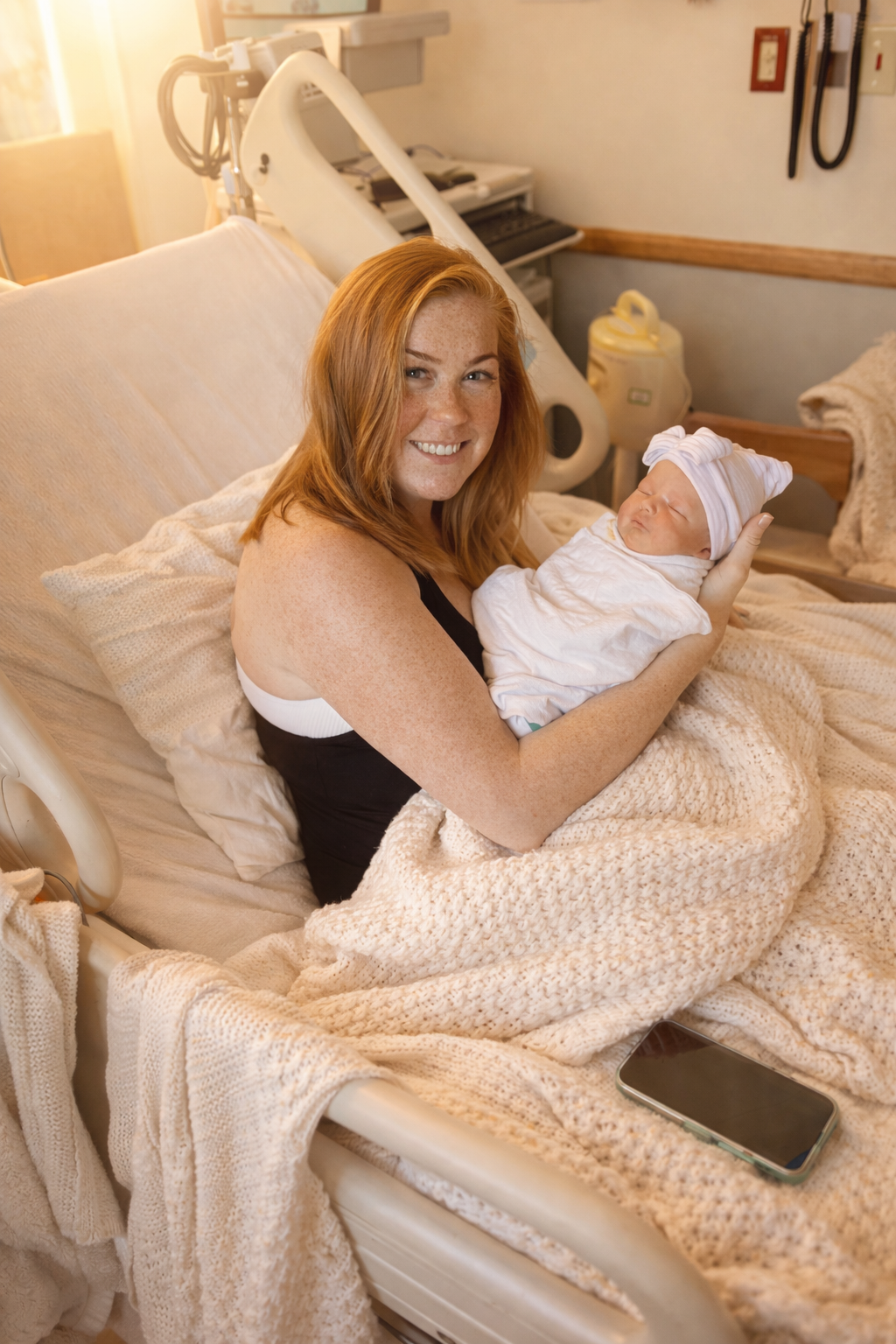 A woman with red hair and freckles smiling and holding a newborn baby in a hospital bed. The baby is wrapped in a blanket and wearing a hat. Medical equipment is visible in the background.