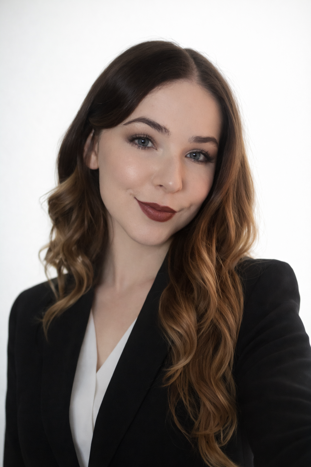 A woman with long wavy brown hair wearing a black blazer and white blouse, smiling at the camera against a plain background.