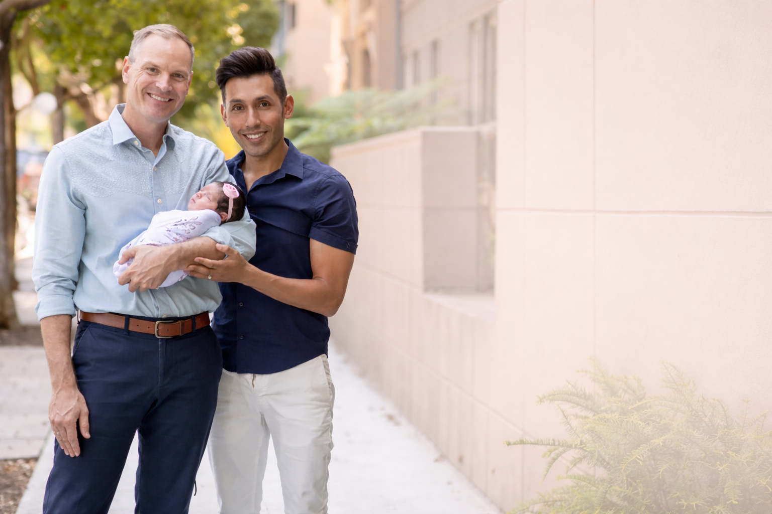 Two men holding a baby girl outdoors on a sidewalk in front of a beige wall and green trees.