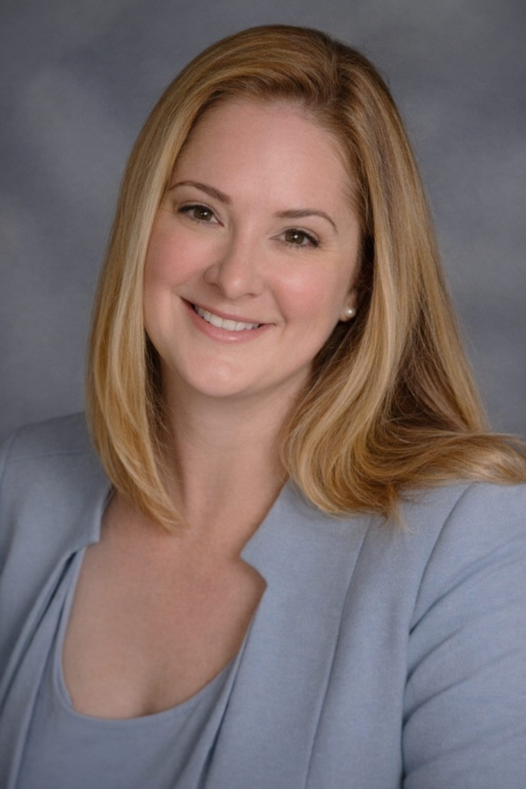 Professional headshot of a smiling woman with shoulder-length red hair, wearing a light gray blazer and pearl earrings, against a gray background.