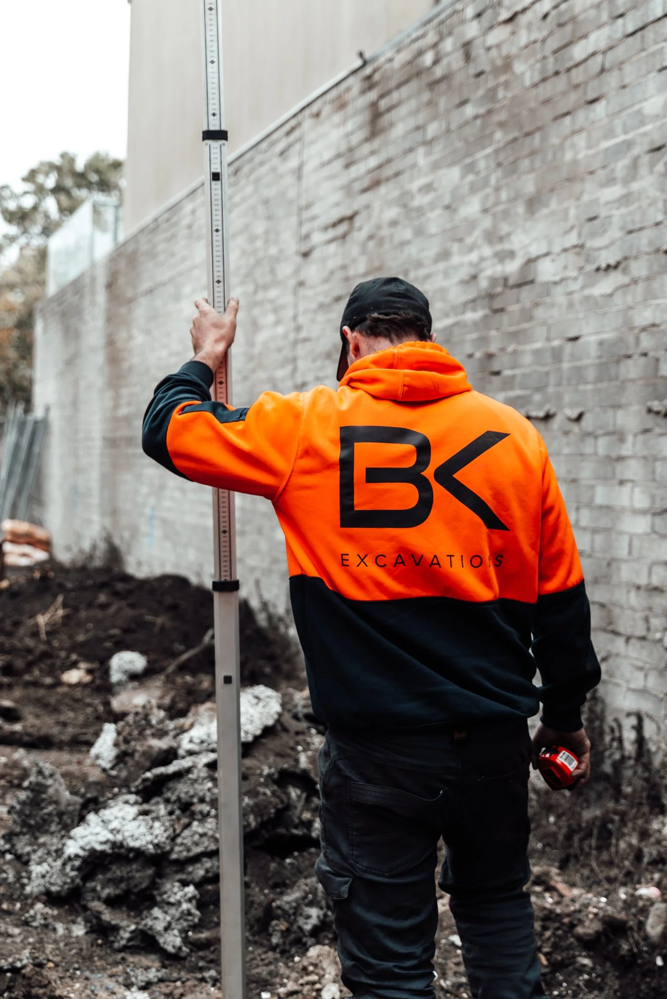 A construction worker in an orange and black jacket with the words 'BK Excavations' on the back, is measuring a brick wall with a height measuring stick. The worker is holding a tape measure in his right hand, and is positioned with his back to the camera. The area appears to be an outdoor construction site with dirt and rocks on the ground, and a brick wall on the right side of the image.