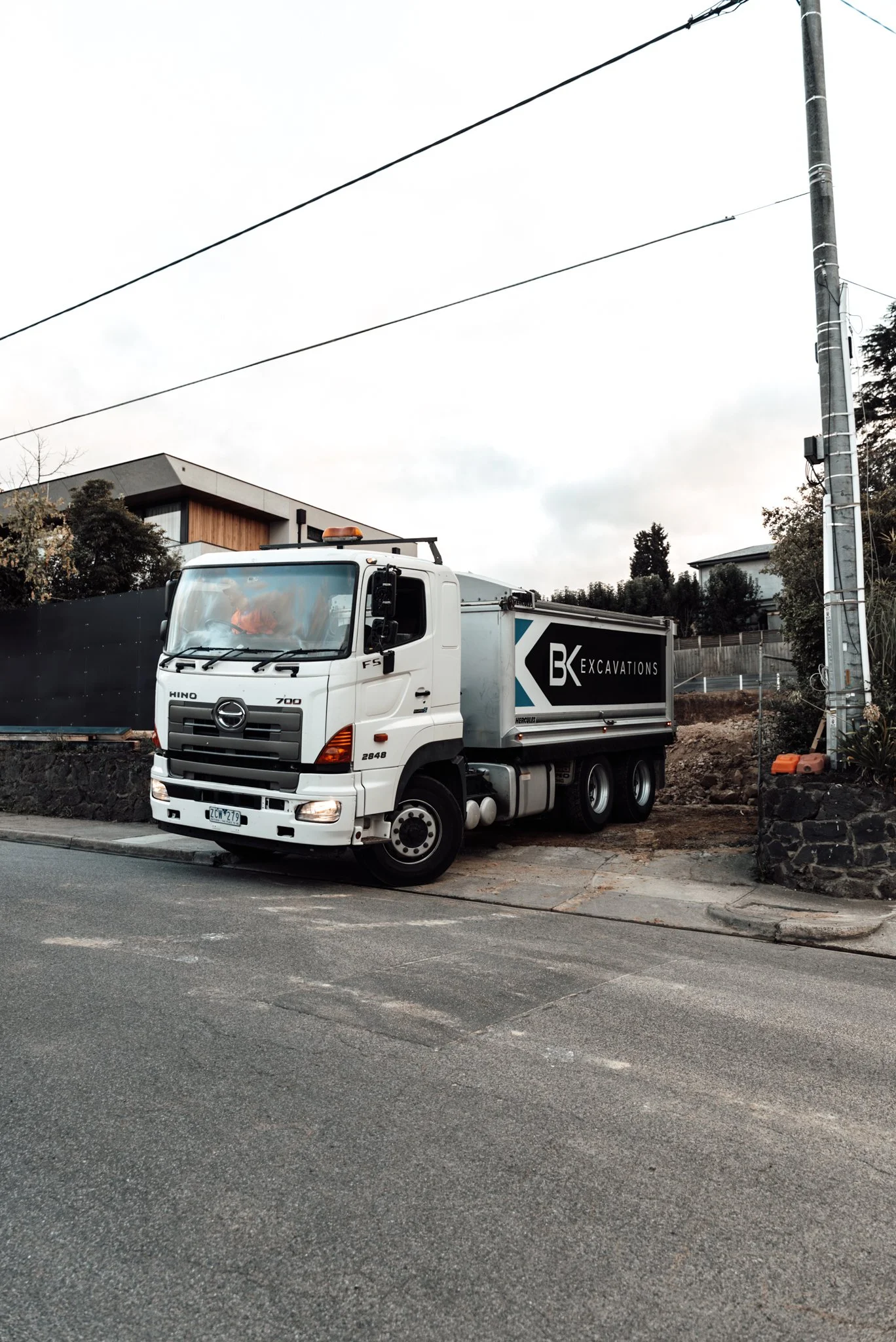 A white Hino 700 truck with a BK Excavations logo on the side, parked beside a residential street with houses and trees in the background. Power lines are visible overhead.