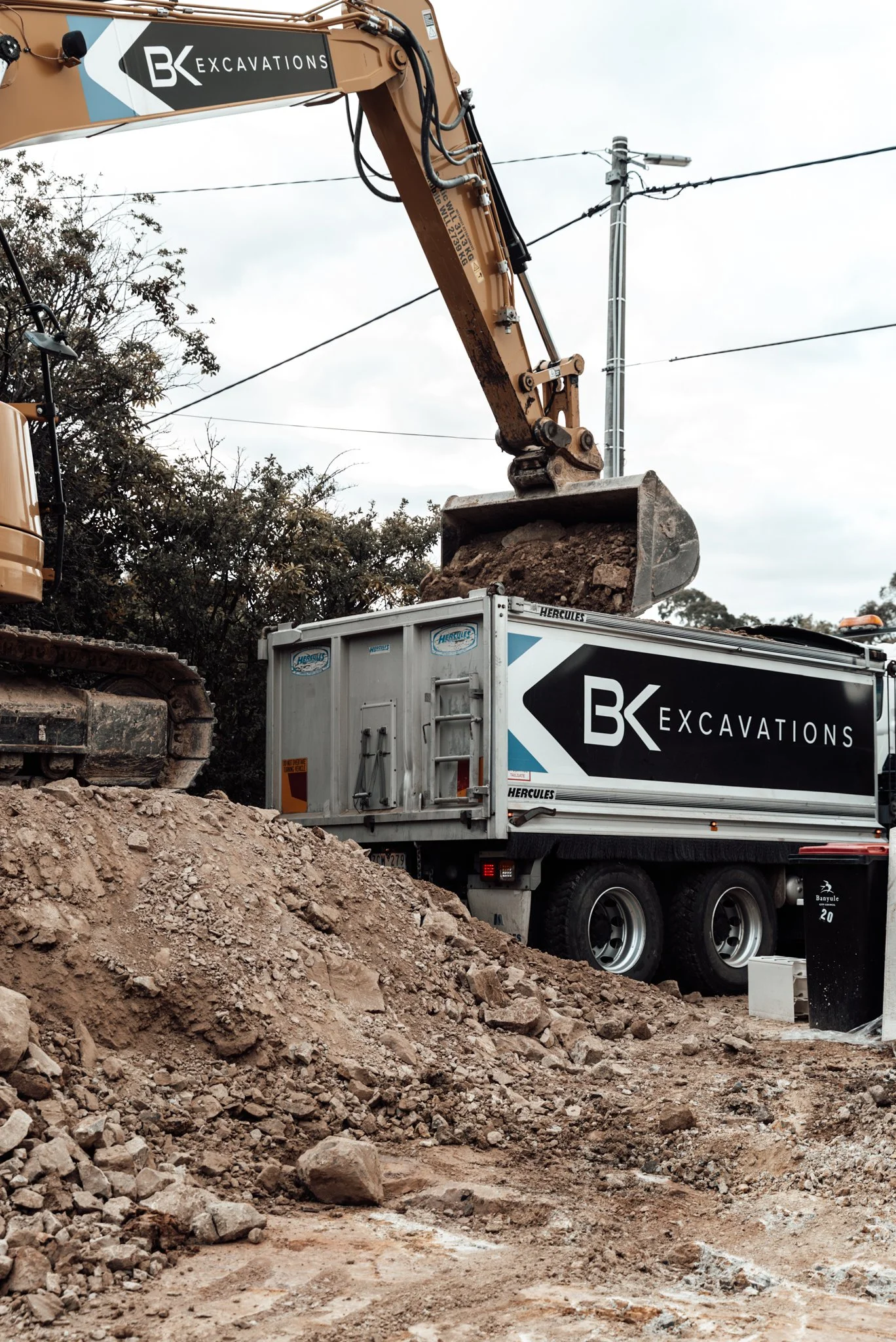 Excavator loading dirt into a dump truck on a construction site, with electrical wires and a pole in the background.