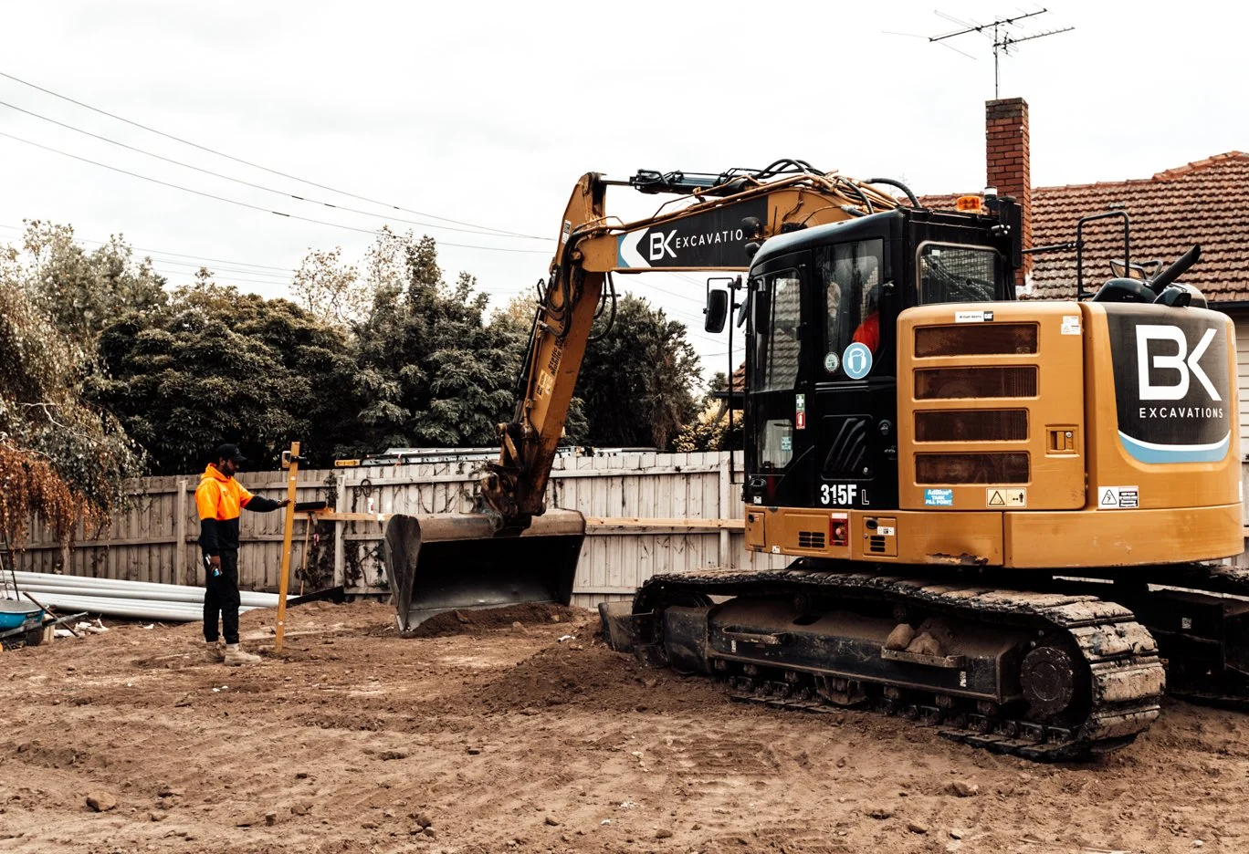A construction worker operating a yellow excavator labeled 'BK Excavations' on a dirt construction site, with a person using a leveling staff nearby and a house with a chimney and tiled roof in the background.