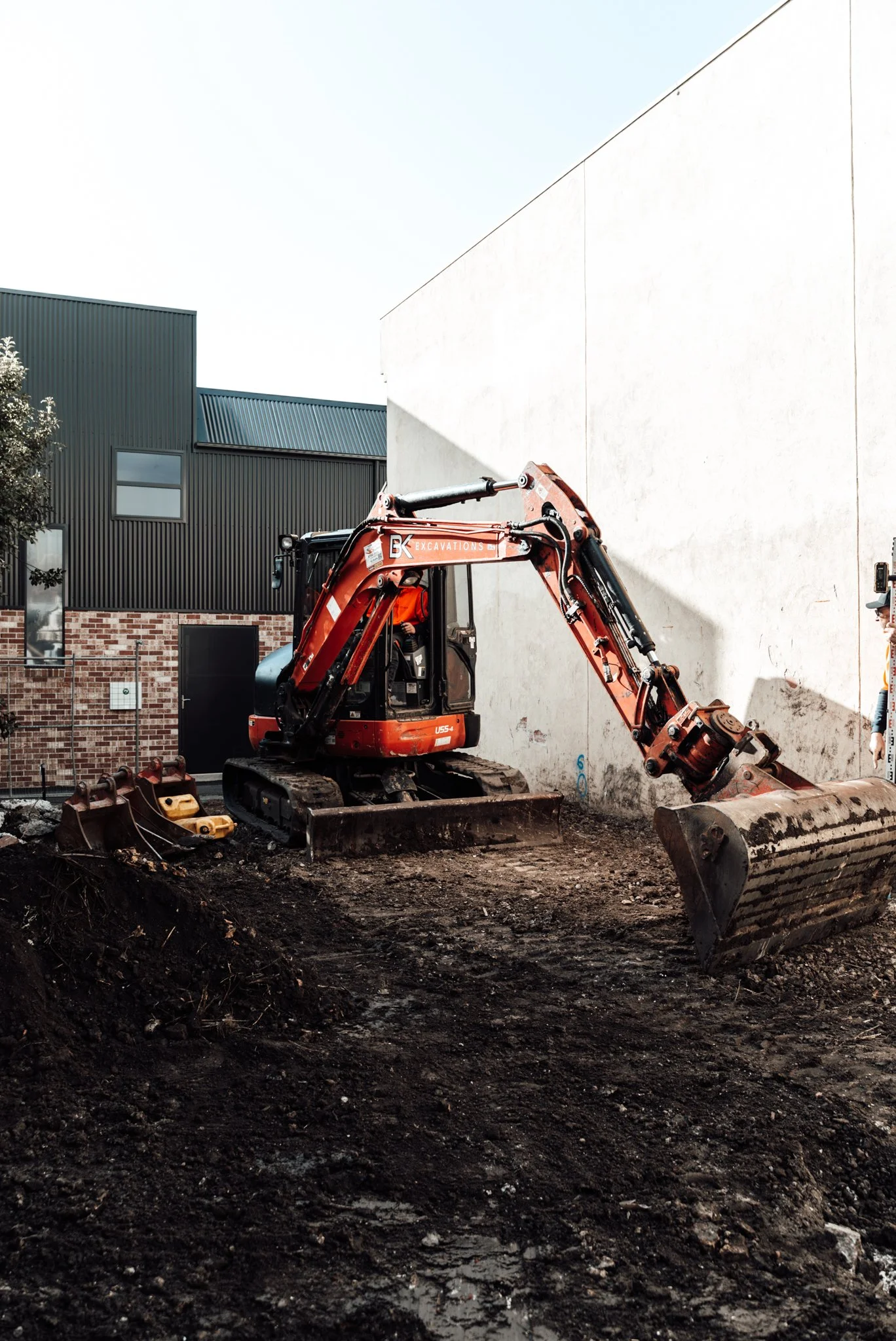 Construction site with an orange excavator digging in dirt, with a modern building featuring dark siding and brickwork in the background.