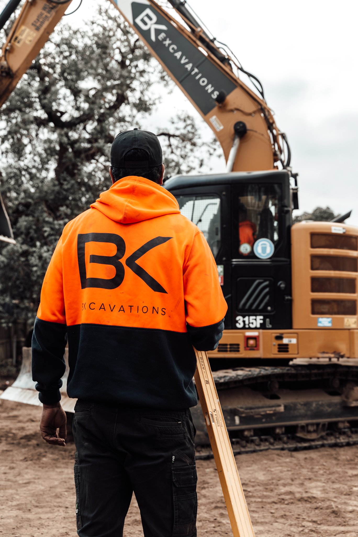 A worker dressed in an orange jacket with the logo 'B K Excavations' on the back, standing in front of a large excavator at a construction site.