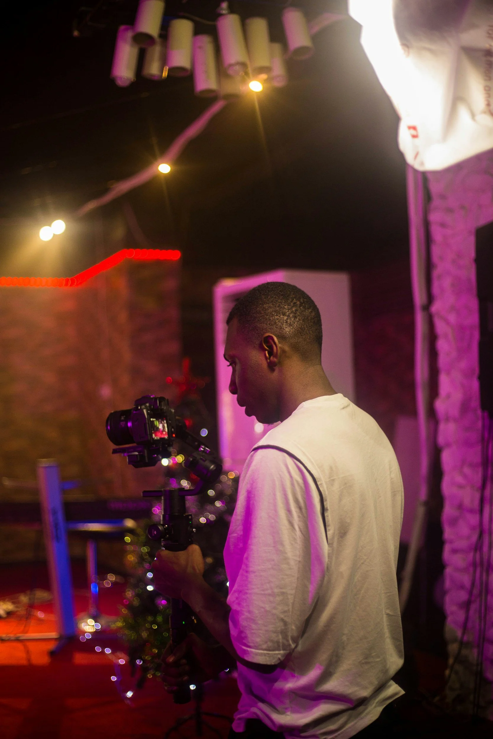 A man holding a camera on a stabilizer in a dimly lit room with colorful lights and a Christmas tree in the background.