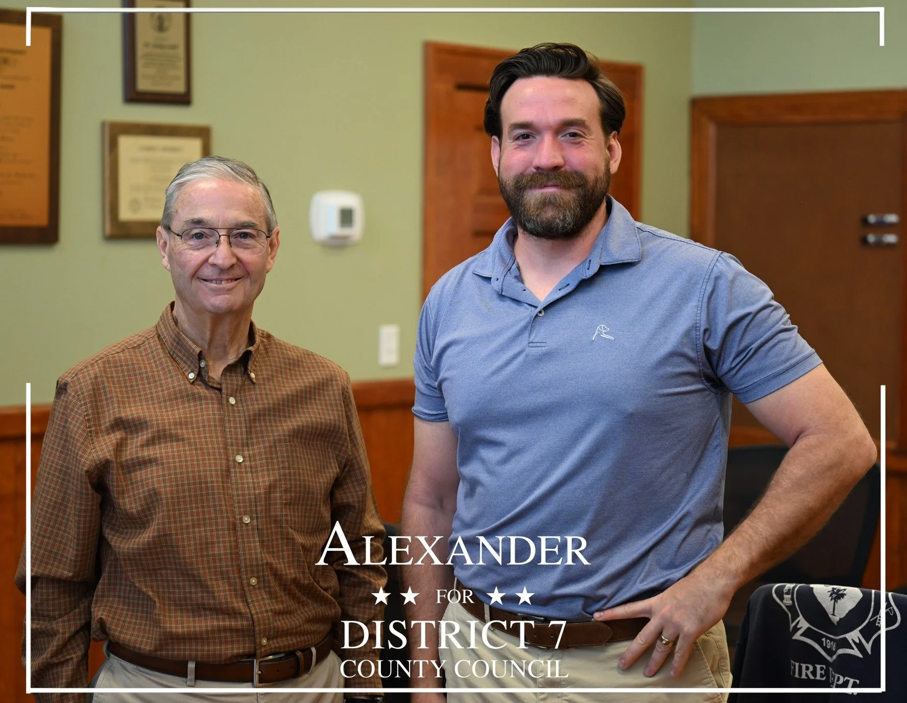 Two men standing inside a room with wood paneling and framed documents on the wall. The older man on the left wears glasses and a brown checkered shirt, smiling. The younger man on the right has a beard, wears a blue polo shirt, and is smiling. Text overlay reads "Alexander for District 7 County Council."
