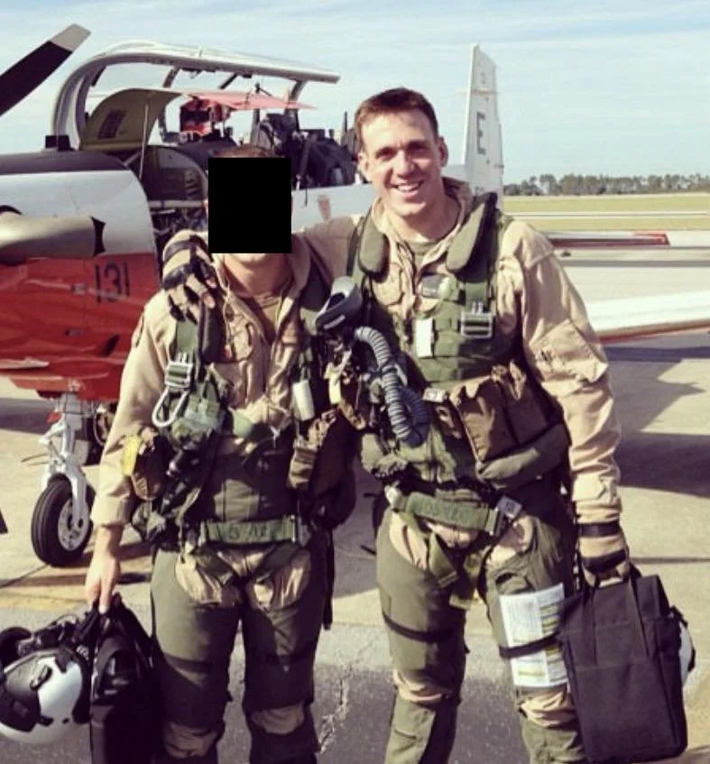 Two military pilots in flight suits standing in front of a military jet on an airfield, smiling at the camera.