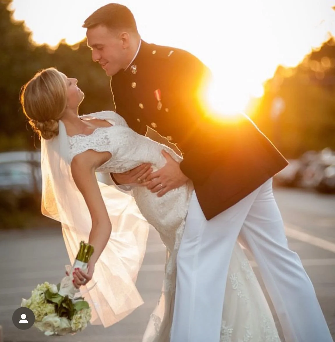 A man in a military uniform holding a woman in a wedding dress during sunset.