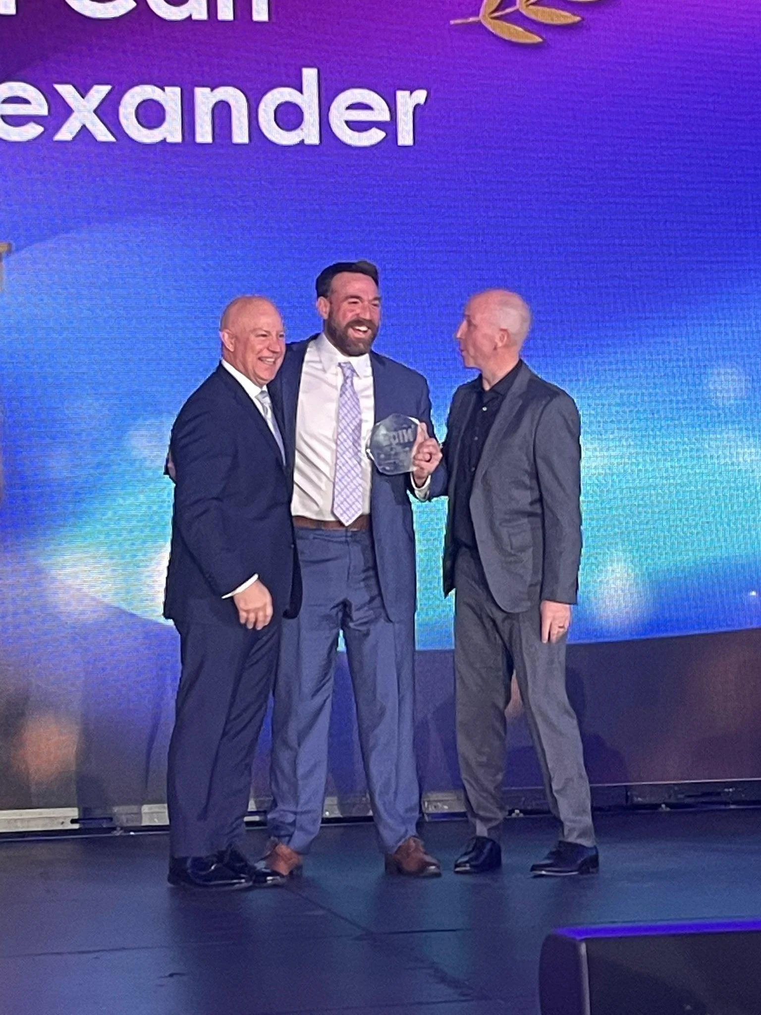 Three men in suits standing on a stage with a purple background, one of whom is holding an award and smiling.