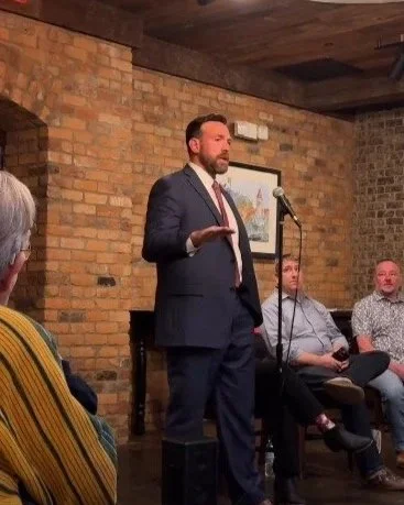 Man in a suit speaking at a microphone in front of a brick wall, with three seated men listening