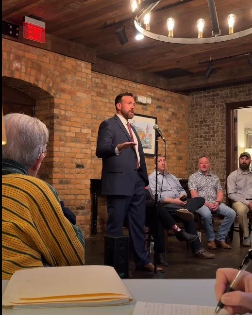 A man in a suit speaking at a microphone during a panel discussion in a brick-walled room, with four seated men listening.