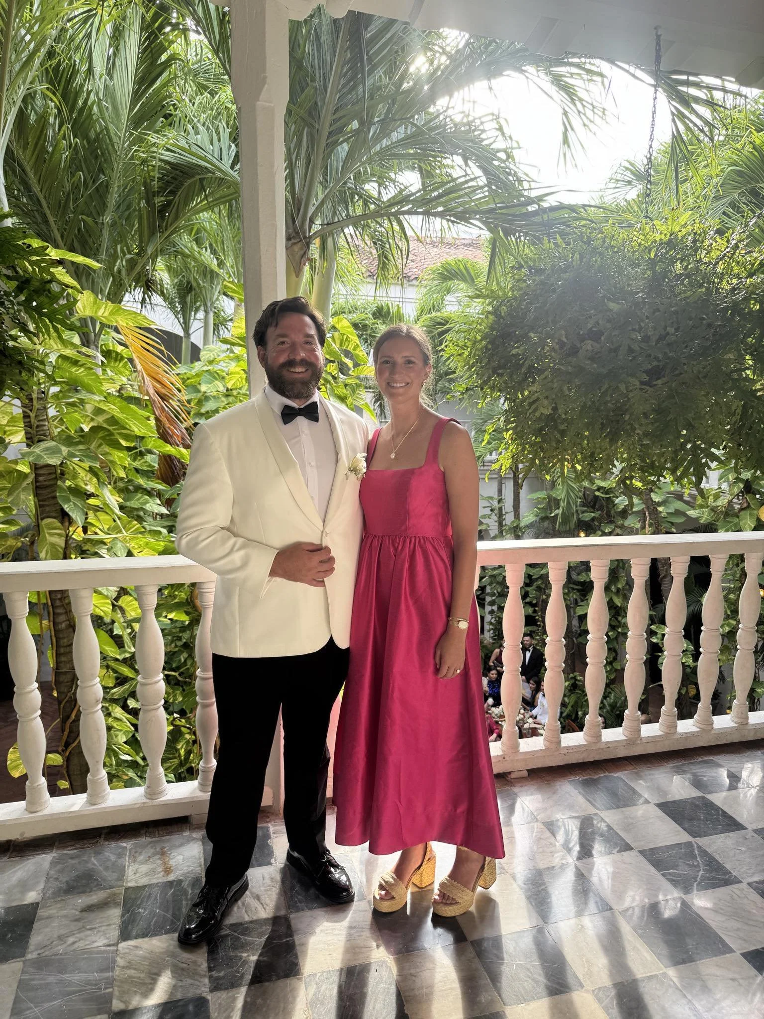 A man in a white tuxedo jacket with a black bowtie and a woman in a pink dress standing on a porch with lush green tropical plants in the background.
