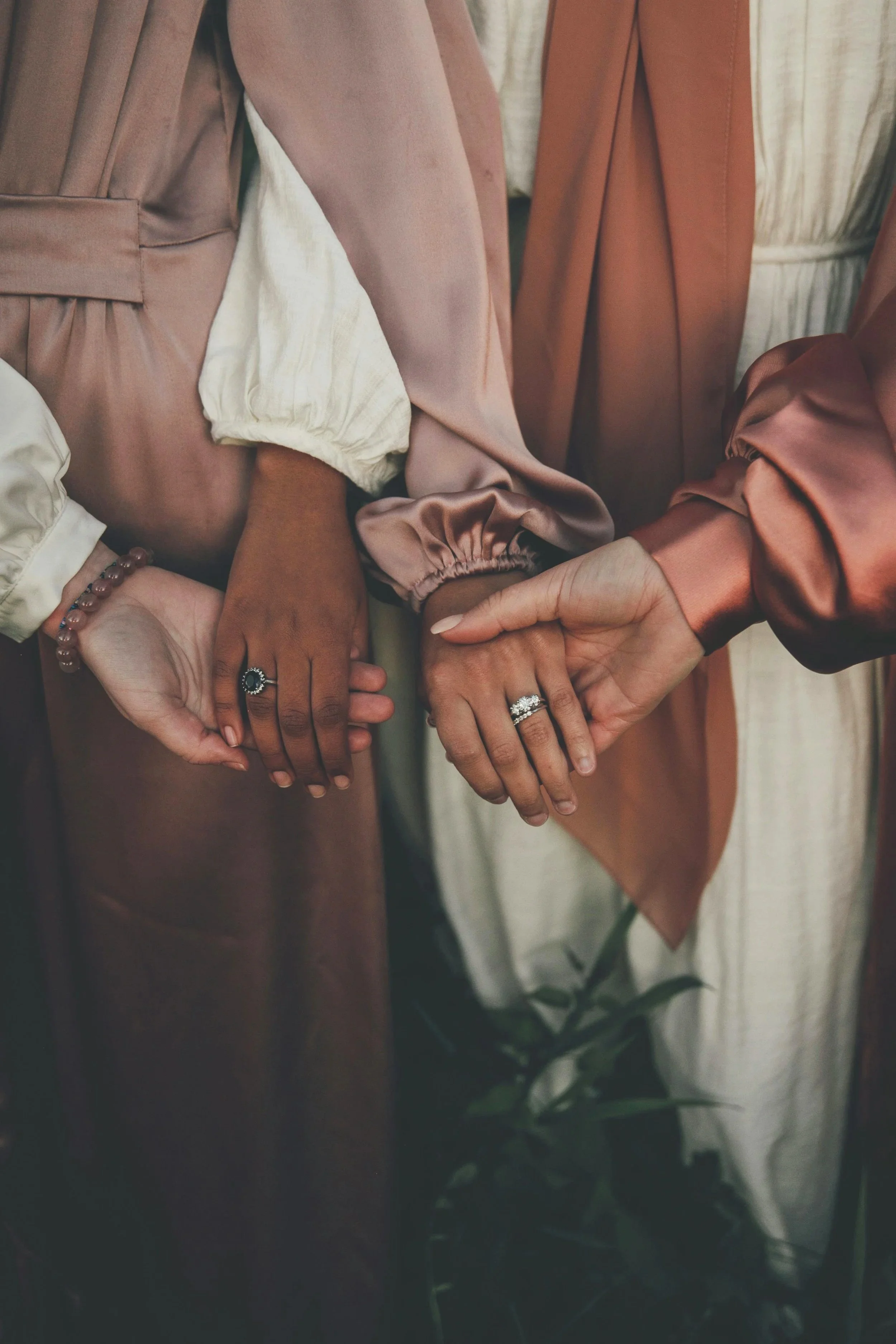 Close-up of three women holding hands, wearing rings and rings, with a background of different satin and fabric coats or dresses, suggesting a wedding or special event.