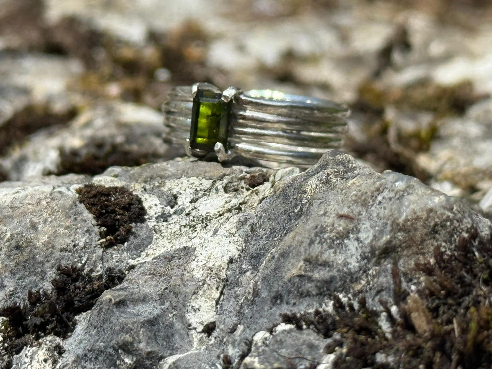 Close-up of a silver ring with a rectangular green gemstone on rough gray and brown rocky ground.