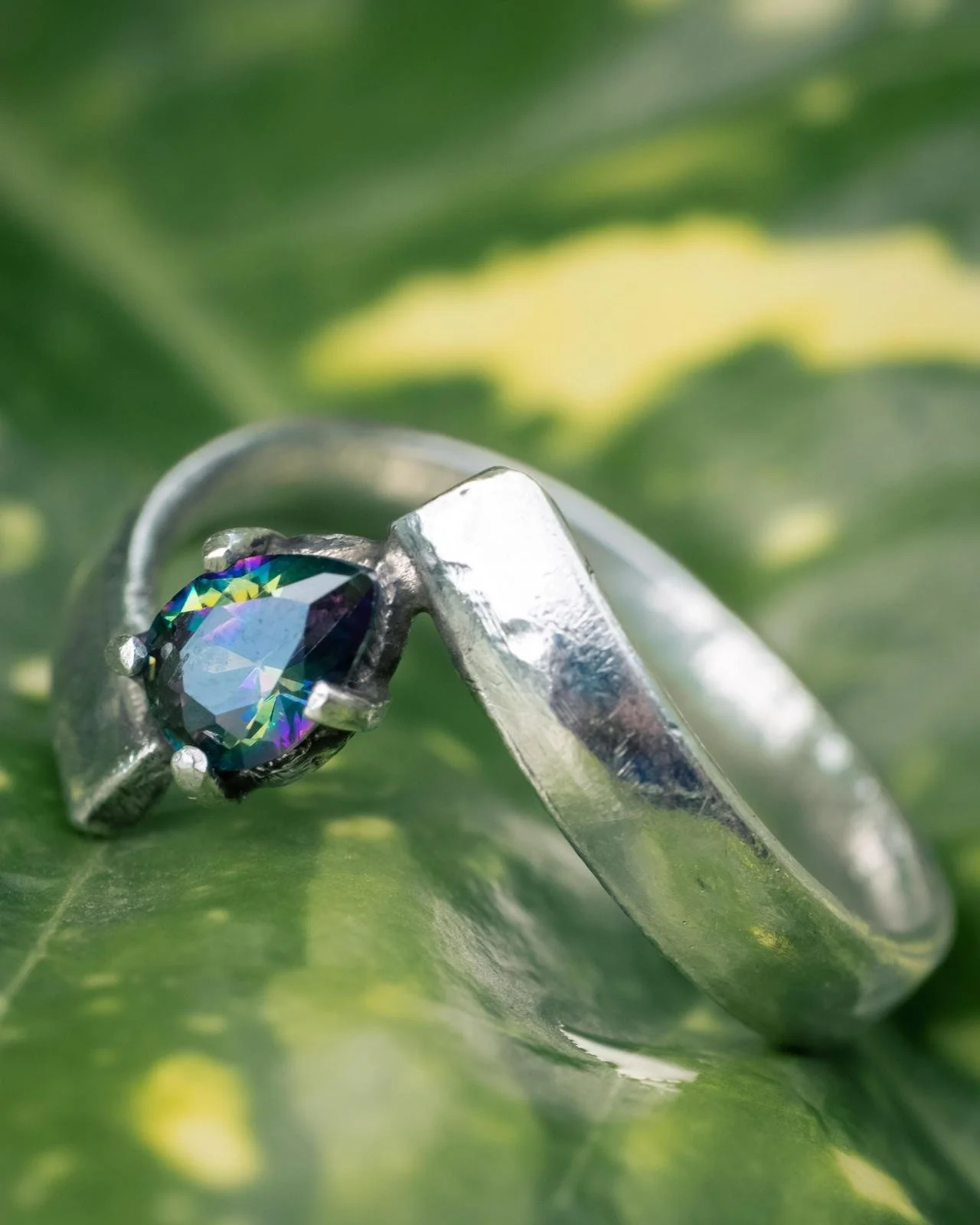 Close-up of a silver ring with a large, multicolored, round-cut gemstone set in a four-prong setting, resting on a green leaf.