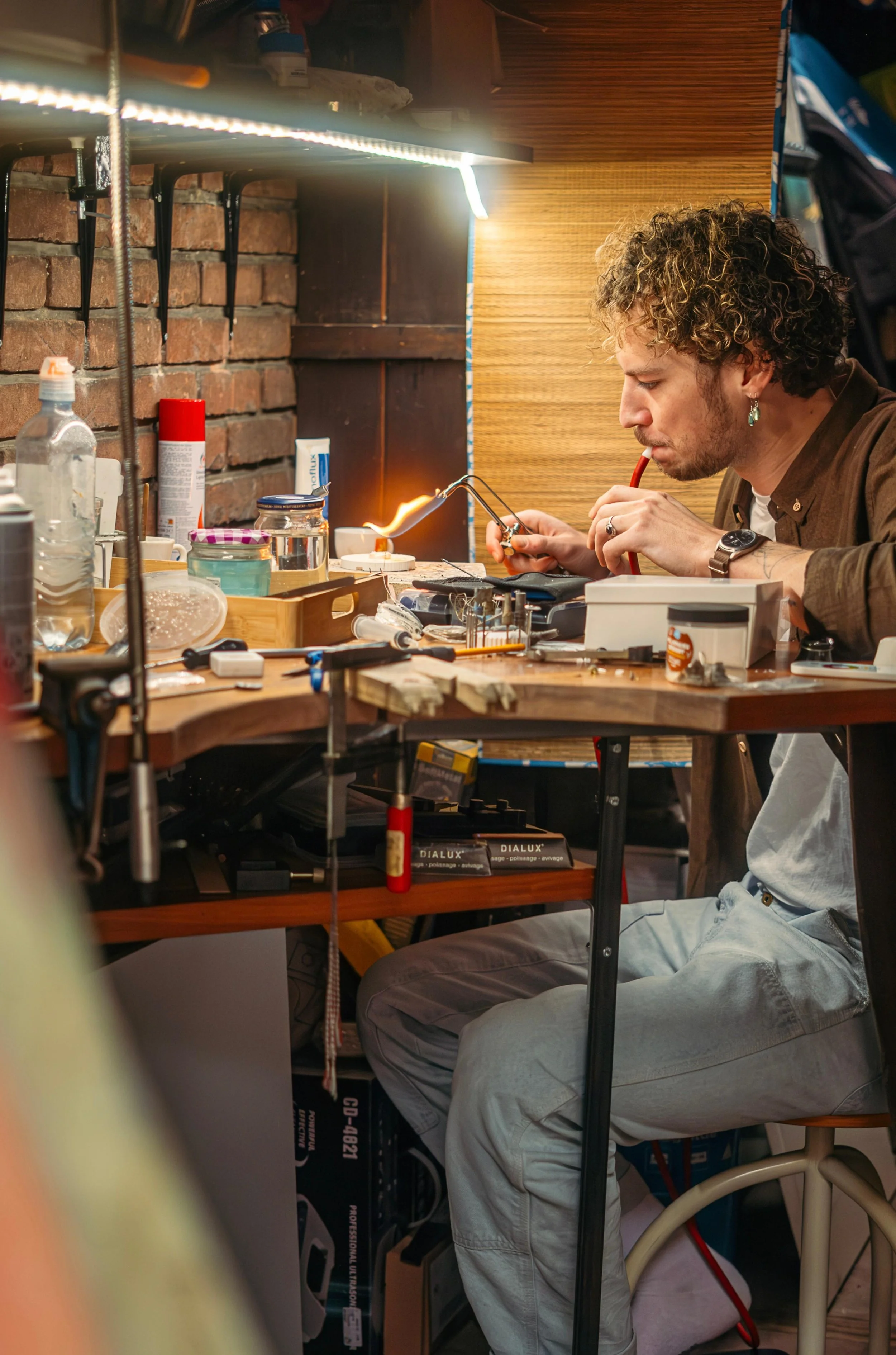 A man with curly hair and earrings peering intently at a small flame while working in a cluttered workshop or lab with various tools and supplies on the table.
