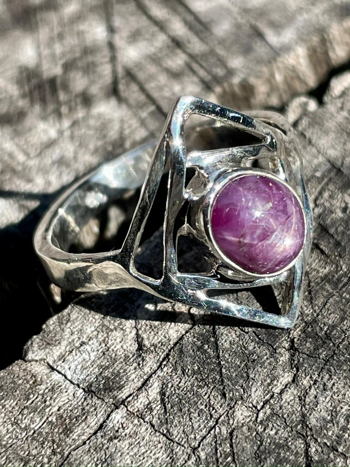 A silver ring with an intricate geometric design and a purple ruby at its center, placed on a textured wooden surface.