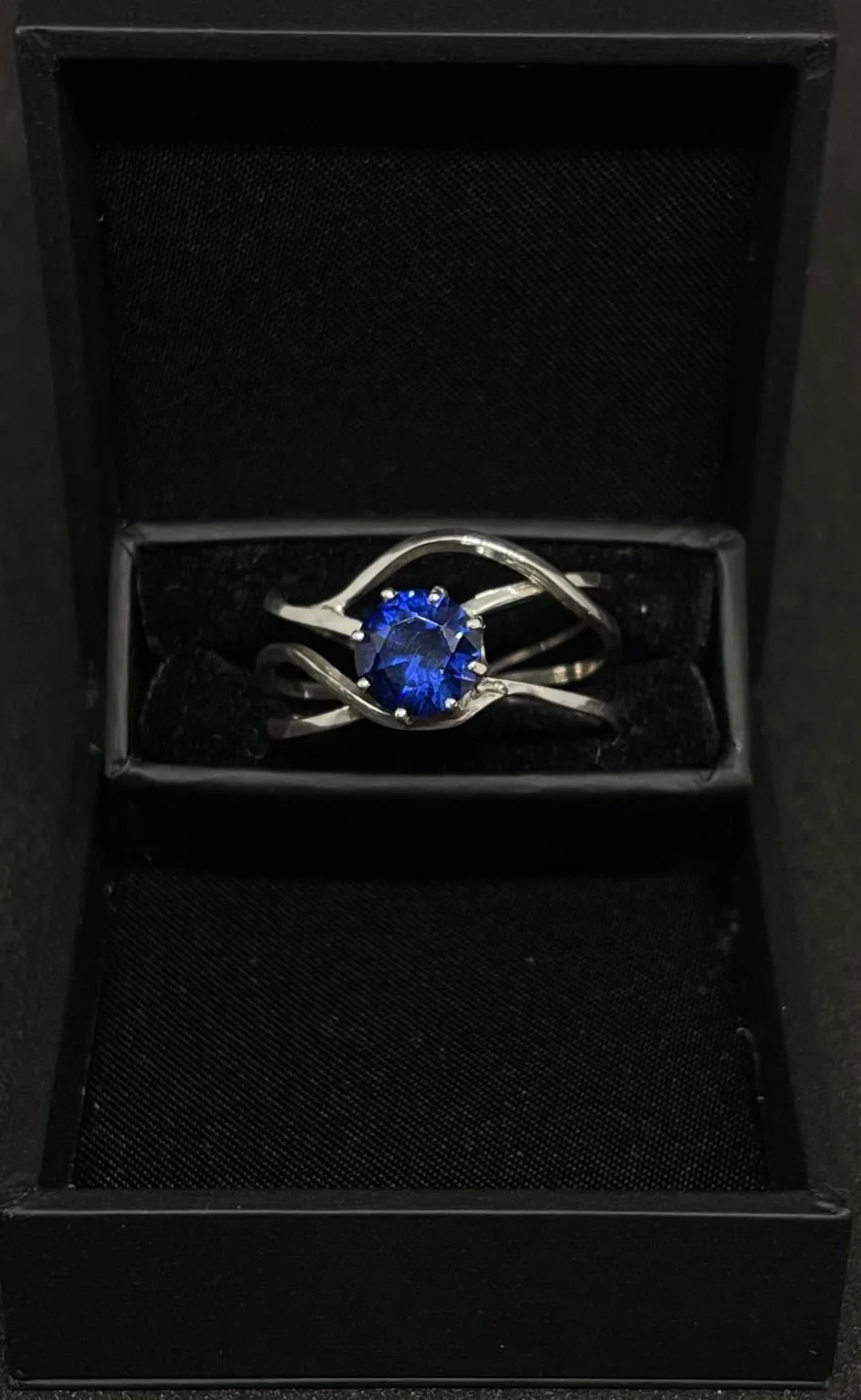 A silver ring with a round blue sapphire in a black jewelry box.
