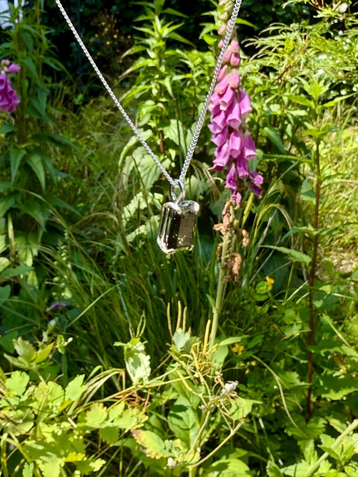 Silver necklace with a rectangular pendant hanging among green plants and purple flowers.