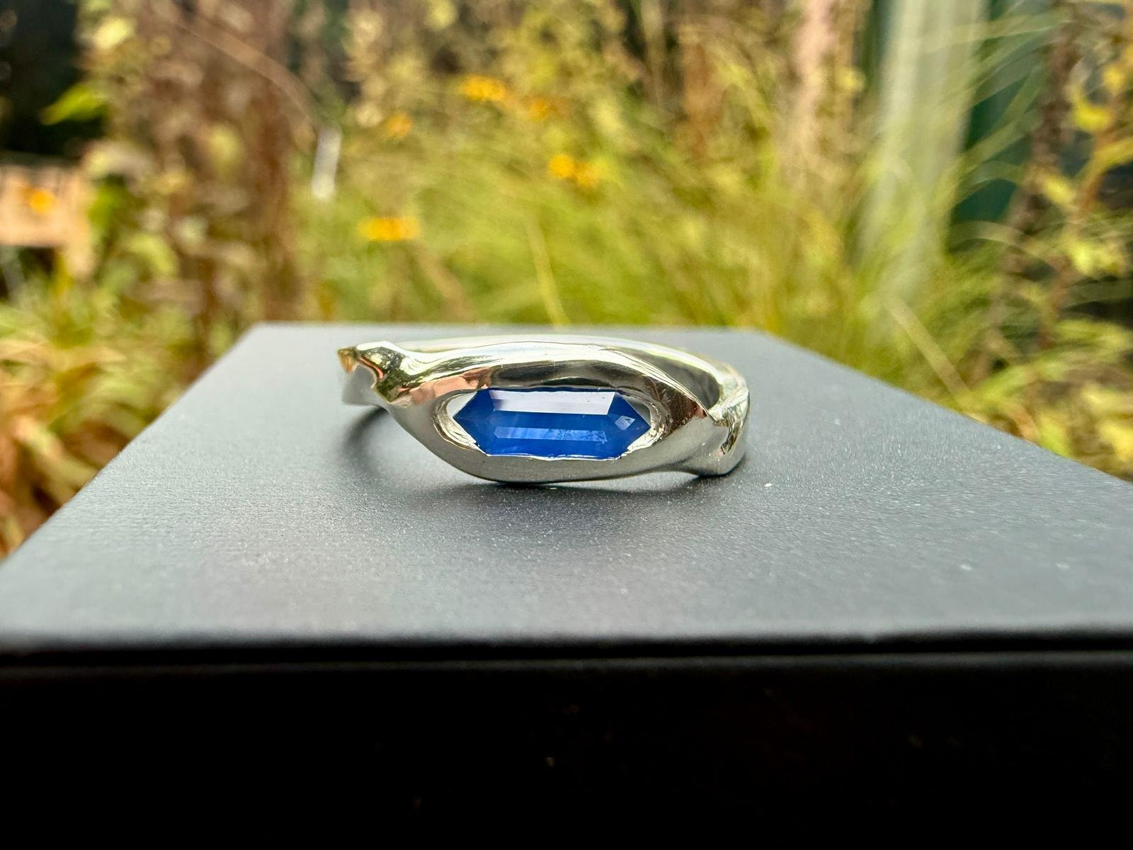 A silver ring with a large, rectangular blue sapphire resting on a black surface outdoors with blurred greenery in the background.