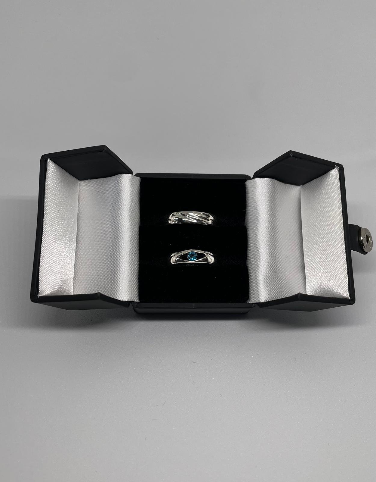 Open black jewelry box with two rings inside, one with a blue gemstone, on a gray surface.