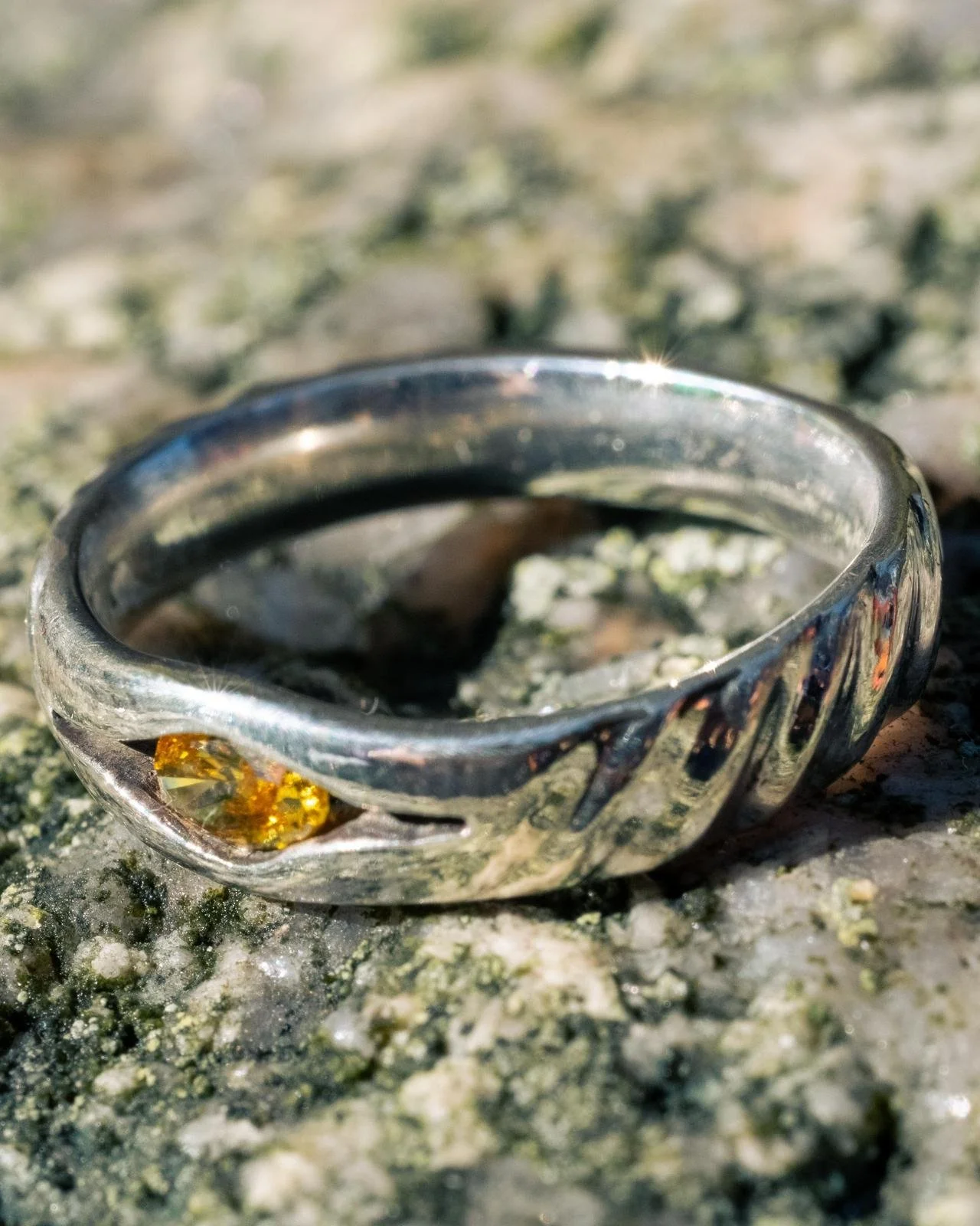 A silver ring with a yellow gemstone on a mossy, rocky surface.