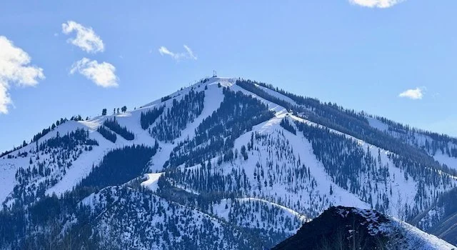 Bald Mountain in Ketchum, Idaho. Snow-covered with ski trails and trees, under a partly cloudy sky.