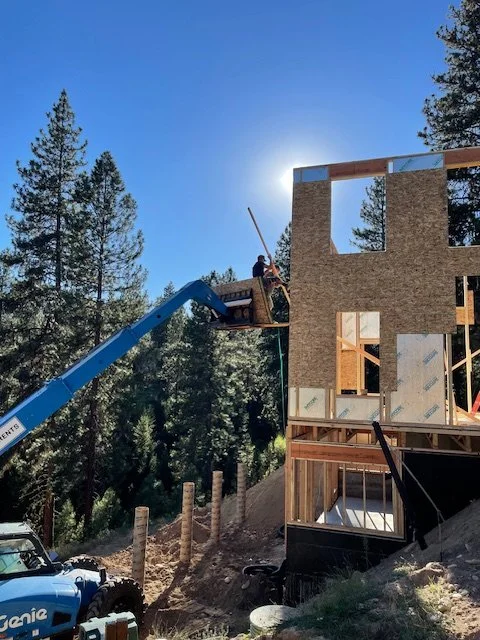 Construction workers installing a large exterior wall on a wooden building frame using a cherry picker, with trees in the background and a clear blue sky.