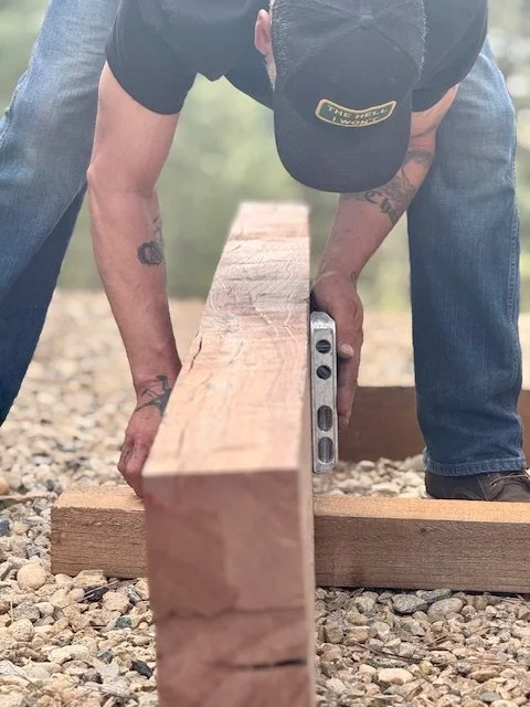Person wearing a baseball cap and jeans using a level to ensure a wooden beam is straight on a gravel surface.