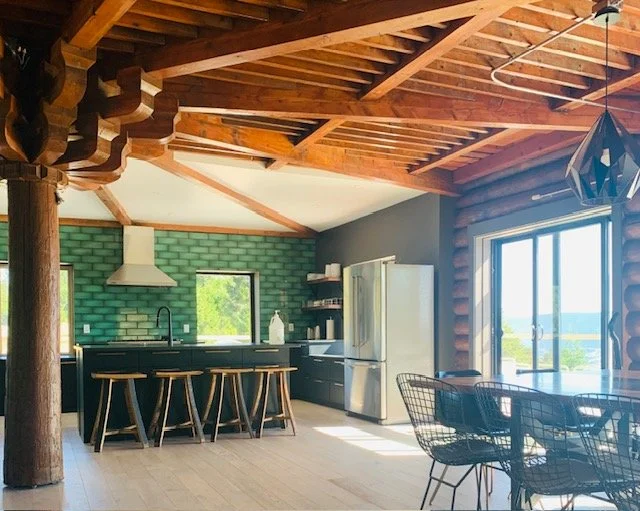Open-concept kitchen and dining area with a log cabin aesthetic, featuring a green tiled backsplash, stainless range hood and refrigerator, black cabinetry, a wooden island with stools, and a glass sliding door leading outside.