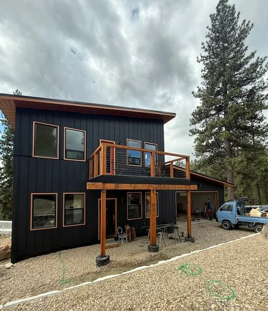A two-story house with dark gray siding, multiple windows, and a wooden balcony on the second floor. The house is located in a wooded area with trees, and there is a gravel driveway with vehicles and outdoor furniture in front.