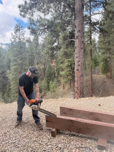 A man wearing a black shirt, jeans, and a baseball cap uses a chainsaw to cut a large wooden beam outdoors on gravel, with a forest of tall pine trees in the background.