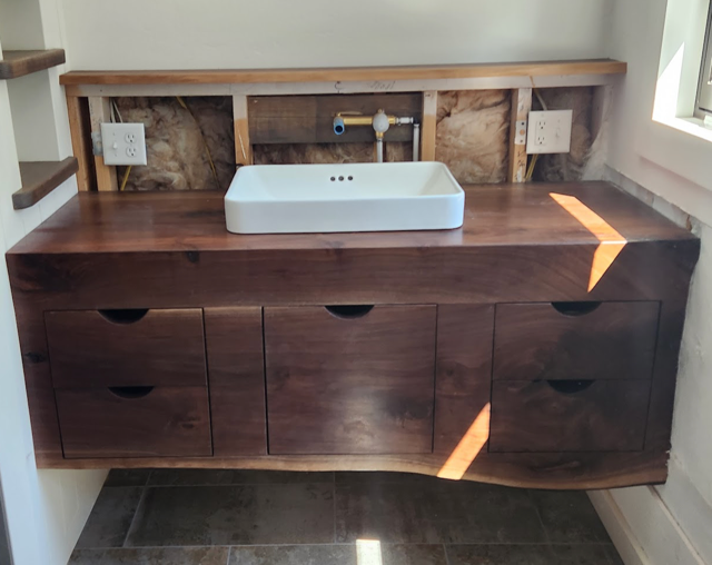 Live edge bathroom vanity with a white rectangular sink, unfinished tile work, and wood grain matched drawers, with visible plumbing and electrical outlets in a partially constructed or remodeled bathroom.