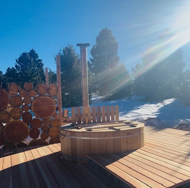 Wood burning hot tub on a deck with snow and trees in the background, sunny sky and sunlight rays